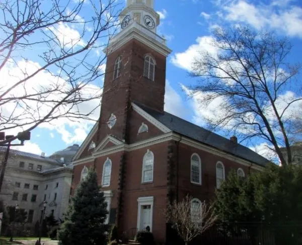 A tall brick church with a white clock‑tower steeple rises above a fenced grassy area with trees under a bright blue sky.