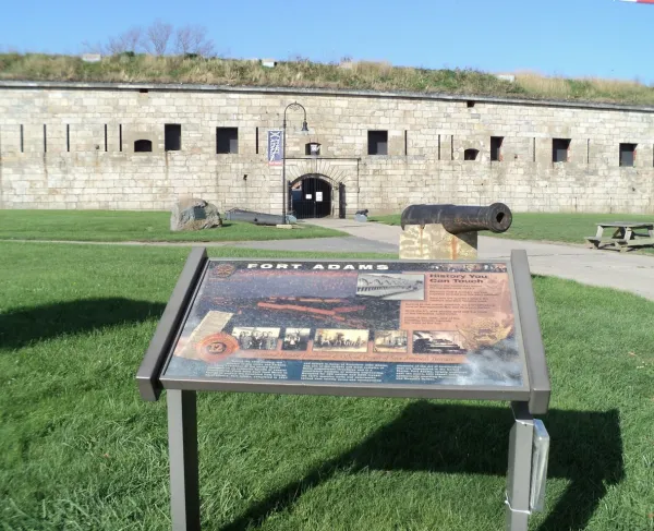A stone fort wall with an arched entrance rises behind a grassy area where an informational sign, an old cannon, and a picnic table sit under a bright blue sky.