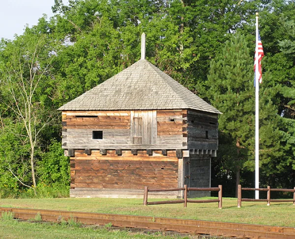 A two‑story wooden blockhouse stands beside a flagpole on a grassy area near dense trees in the background.