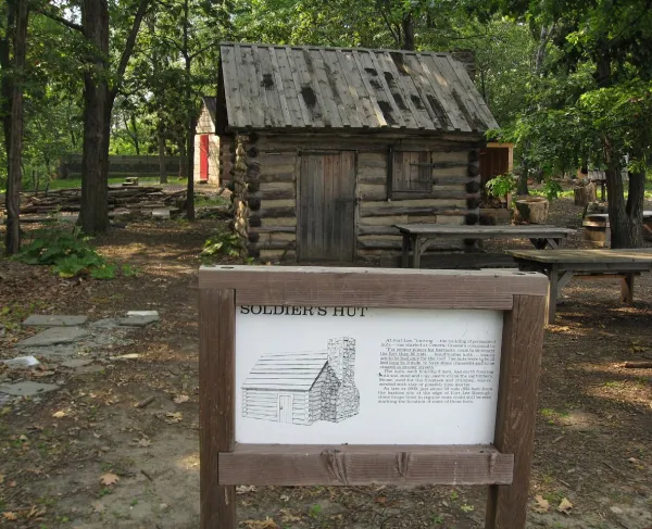 A small reconstructed Revolutionary War–era log cabin stands in a wooded clearing. The structure is labeled “Soldier’s Hut” on an informational sign in front of it. 