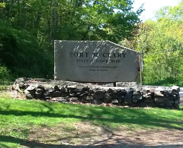  A stone sign reading “Fort McCleary State Historic Site” stands beside a road, surrounded by a low stone wall, grass, and trees.