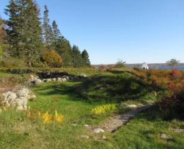 A grassy stone‑lined path curves toward a distant blue body of water and lighthouse, bordered by evergreen trees and autumn foliage under a clear sky.