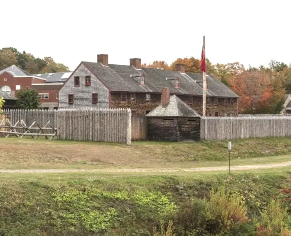 A colonial-era fort enclosed by a tall wooden palisade stands amid autumn-colored trees, with weathered timber buildings, a flagpole, and grassy grounds viewed from across a shallow ditch.