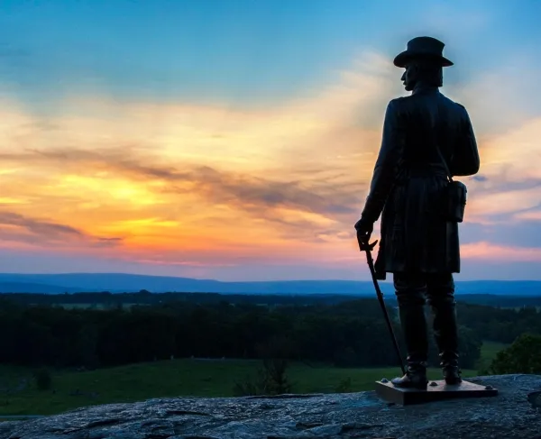 A statue stands at Little Round Top against a vibrant sunset.
