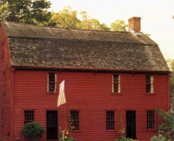 A red wooden colonial‑style house with a steep shingled roof and small‑paned windows stands amid green grass and trees, with an American flag mounted near one of its front doors.