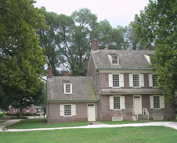 A brick colonial‑style house with cream shutters and multiple chimneys stands amid green grass and tall trees.