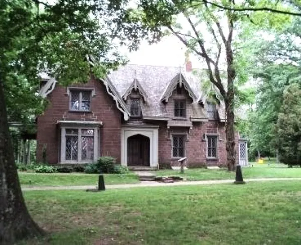 A large brick Gothic‑style house with steep gables and tall narrow windows stands among mature trees on a grassy lawn.