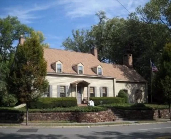 A tan colonial‑style house with dormer windows, brick chimneys, and a columned porch stands behind trimmed hedges and a stone retaining wall, with an American flag flying beside it.