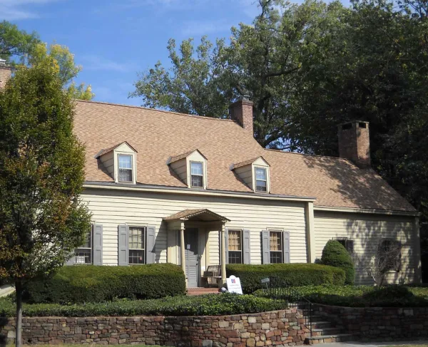 A tan colonial‑style house with dormer windows, brick chimneys, and a columned porch stands behind trimmed hedges and a stone retaining wall, with an American flag flying beside it.