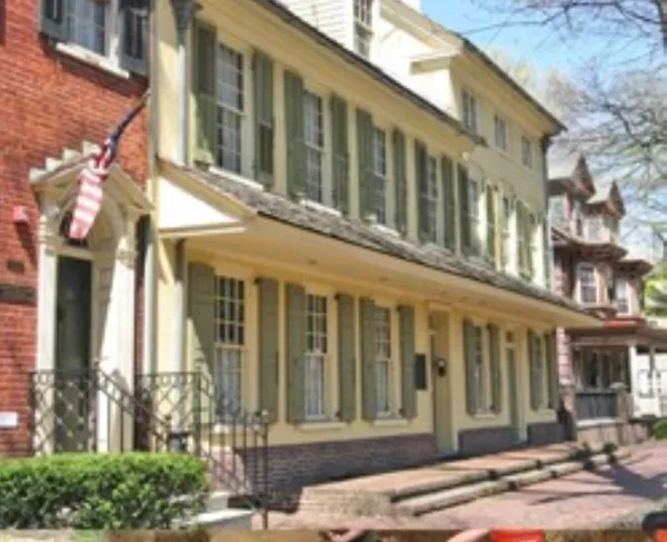 A long two‑story cream-colored historic building with green shutters, stands along a brick sidewalk. An American flag hangs beside the entrance, and neighboring historic buildings line the street.