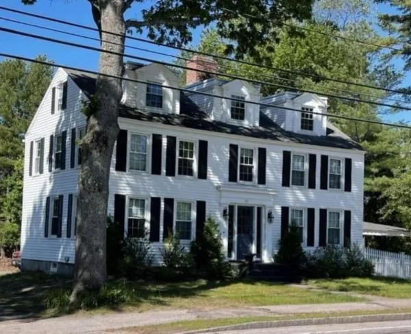 A large white colonial‑style house with black shutters and three dormer windows stands behind a picket fence, surrounded by trees along a roadside.