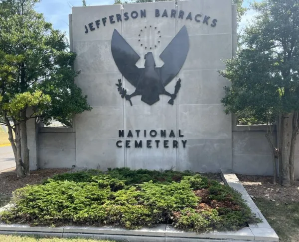 A stone entrance sign for Jefferson Barracks National Cemetery stands among neatly trimmed shrubs and trees. The sign features a carved emblem of an eagle with outstretched wings holding an olive branch and arrows, with stars above its head.