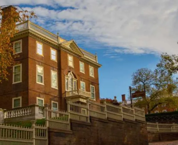 A large brick building with white trim and classical details stands behind a tall white fence, framed by autumn trees under a partly cloudy sky.