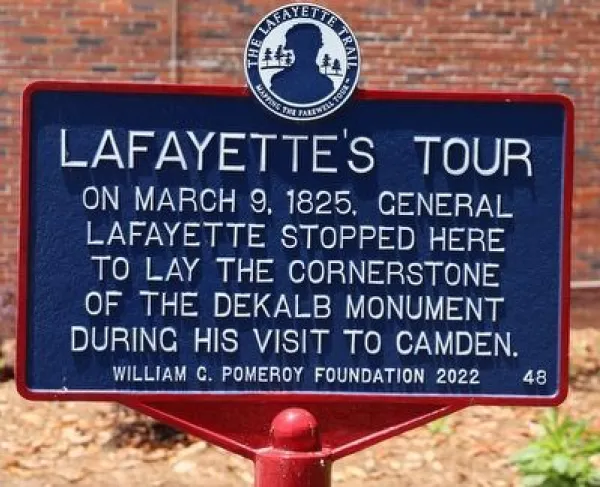 A blue historical marker titled “Lafayette’s Tour” stands in front of a brick wall and plants, describing General Lafayette’s 1825 visit to lay the cornerstone of the DeKalb Monument in Camden.
