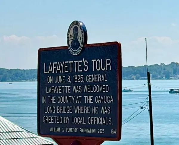 A blue historical marker outlined in red with white lettering titled “Lafayette’s Tour” stands on a lawn in front of a lake commemorating Lafayette's visit to this city.