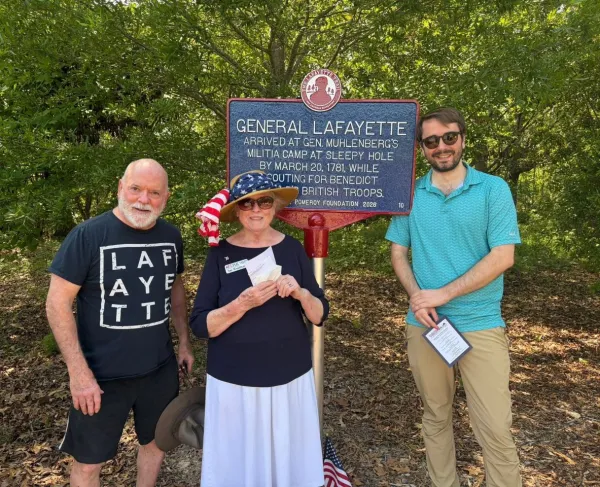 A blue historical marker outlined in red with white lettering titled “General Lafayette” stands on a lawn in front of trees and 3 people commemorating Lafayette's encampment in this city.