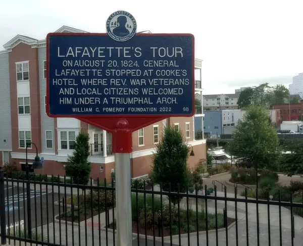 A blue historical marker on a red post describes General Lafayette’s 1824 visit, standing in front of modern brick and white buildings with a fenced walkway and landscaped greenery.
