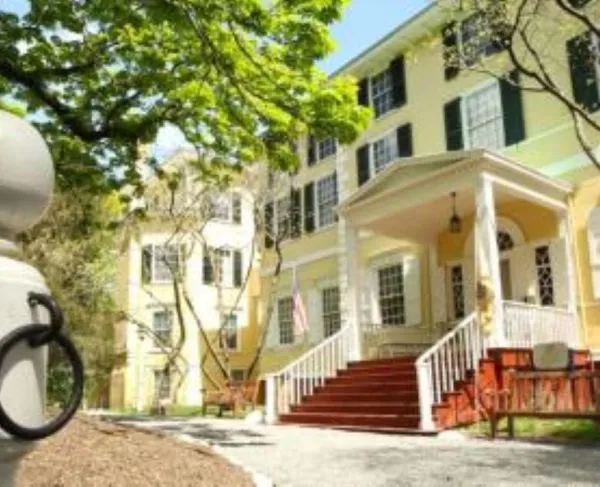 A large yellow historic house with white trim and a columned front porch stands among trees on a sunny day.
