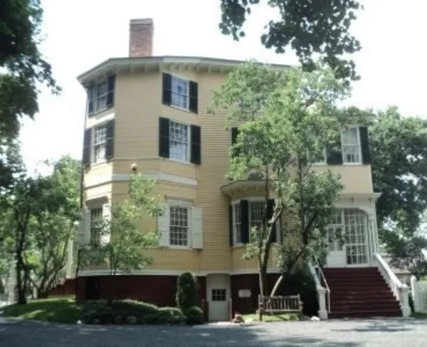 A large light‑yellow historic house with white trim, dark shutters, and an octagonal shape stands among trees with a brick foundation and a prominent front staircase.