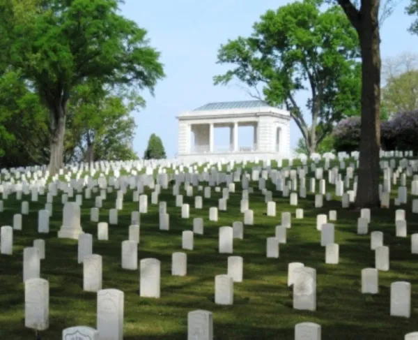 Rows of white headstones line a grassy hillside in a quiet cemetery. Behind them stands a white stone memorial building with columns and a metal roof, surrounded by tall green trees that create a calm, shaded atmosphere.