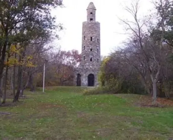 A tall cylindrical stone tower with small windows and an arched entrance stands in a grassy clearing surrounded by trees showing early autumn color.