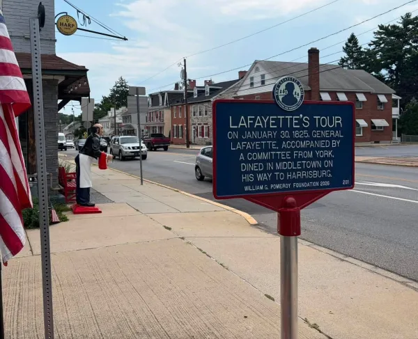 Close up of a sign that contains a description of Lafayette's Tour with a street lined with buildings behind it.