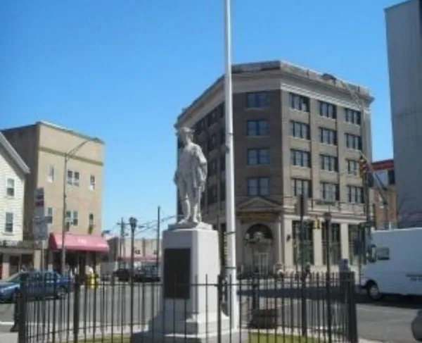A colonial era militia statue enclosed by a circular black fence stands in a small plaza with an American flag behind it, surrounded by multi‑story city buildings under a bright blue sky.