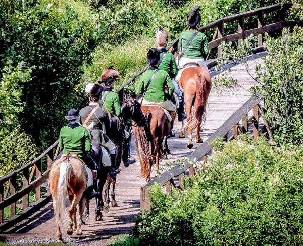 Five riders in matching green jackets and black hats cross a wooden bridge on horseback through dense green foliage.