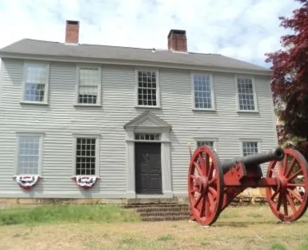 Colonial era 2-story manor house with 9 windows trimmed in white, with 2 chimneys and a red cannon in the front yard.