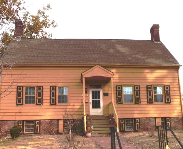 A two‑story yellow colonial house with dark brown trim and twin brick chimneys stands behind a small front porch and brick walkway.