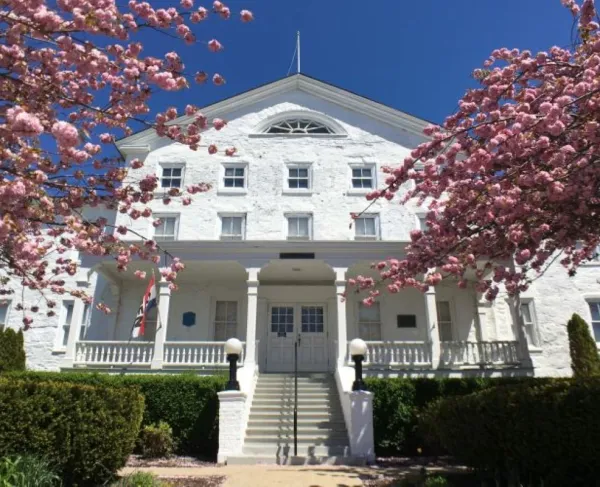 A white colonial‑style building with a central staircase and double doors is framed by bright pink flowering trees, with an American flag near the entrance and a clear blue sky overhead.