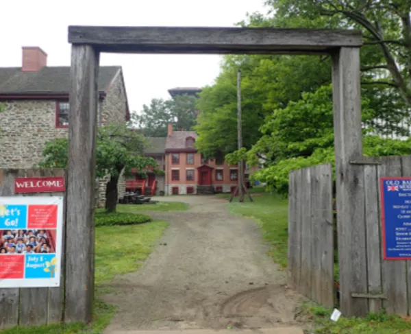 A wooden entrance gate stands at the Old Barracks Museum, built from two tall posts and a horizontal beam.  Beyond the gate, a gravel path leads to historic stone and brick buildings with red trim, surrounded by trees and greenery.