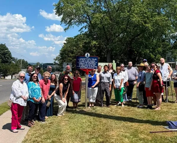A blue historical marker outlined in red with white lettering titled “Lafayette’s Tour” stands on a lawn in front of large tree with a group of peoople commemorating Lafayette's visit to this city.