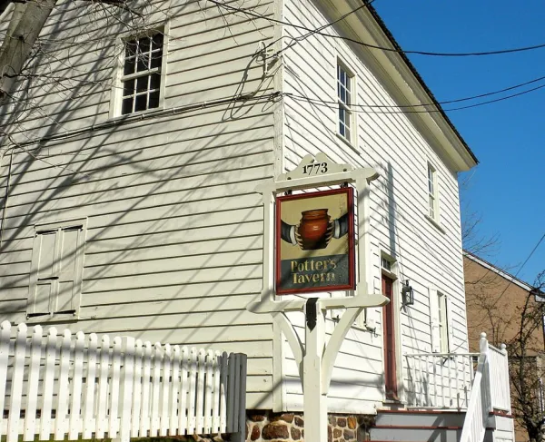 A white historic salt box style building with a stone foundation and an exterior stairway stands behind a sign reading “Potter’s Tavern 1773” topped with a brown pot illustration.