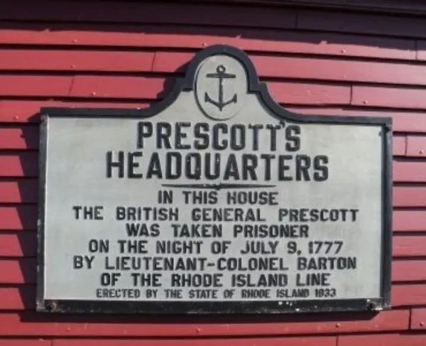 A rectangular historical marker to British General Prescott mounted on a red wooden wall.