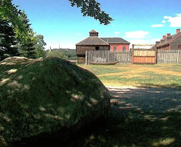A large boulder sits in the shade before a reconstructed colonial-era fort enclosed by a wooden palisade fence, with historic buildings and open grounds under a bright blue sky.