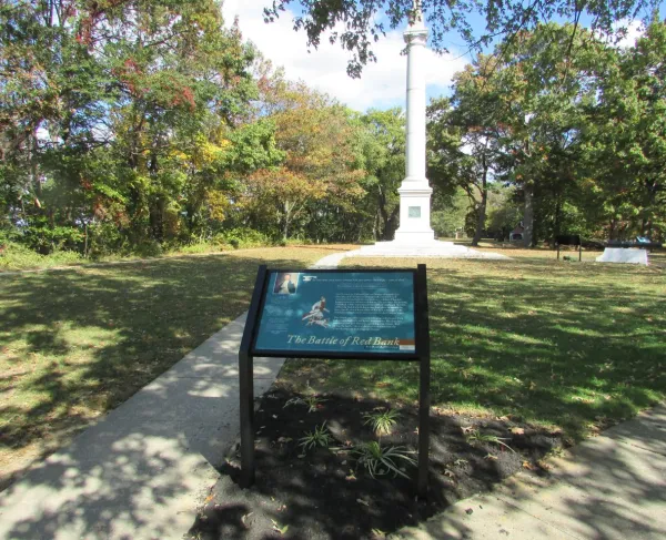 A tall white monument stands behind an informational sign about the Battle of Red Bank in a grassy area with autumn trees.