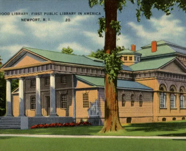 A classical brick building with a columned portico and triangular pediment stands behind a small lamp post, surrounded by trees and greenery.