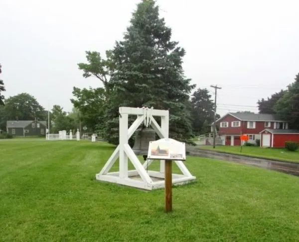 A historic bell hangs in a white wooden frame on a grassy village green, with a large evergreen tree behind it and red houses lining a quiet road.