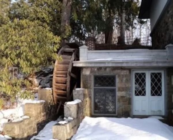 A rustic stone mill with a large wooden water wheel stands in the snow among evergreen trees.