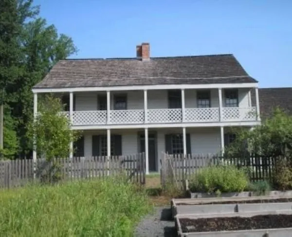 A two‑story wooden house with a long lattice‑railed balcony stands behind a fenced yard with raised garden beds and surrounding trees.