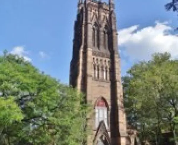 A tall brown stone Gothic‑style church tower with pointed arches and ornate spires rises above trees under a bright blue sky.