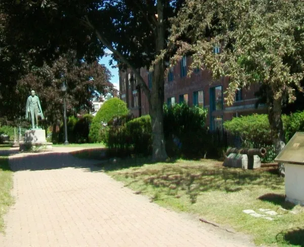A brick pathway shaded by mature trees leads through a historic park featuring a bronze statue, an interpretive marker, and a small cannon displayed beside a red-brick building on a sunny day.