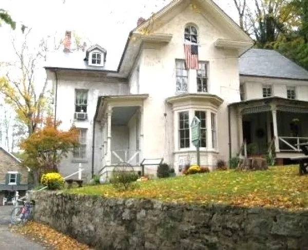 A large light‑colored house with multiple gables, a stone wall in front, and an American flag above the porch sits among autumn trees with fallen leaves.