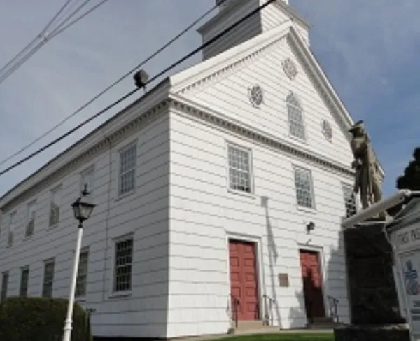 A white wooden church with a tall clock‑tower steeple and red doors stands behind a stone‑bordered lawn with a lamppost and statue of a Revolutionary war soldier.