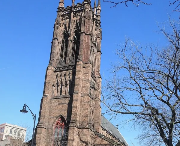 A tall brown stone Gothic‑style church tower with pointed arches and ornate spires rises above trees under a bright blue sky.