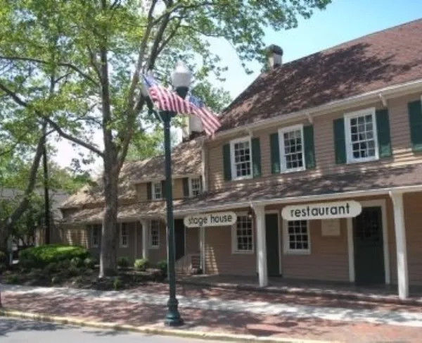 A tan historic building with green shutters and a columned porch stands along a brick sidewalk, shaded by large trees and marked by a streetlamp flying an American flag.
