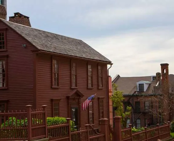 A reddish‑brown historic wooden house with tall narrow windows and an American flag at the entrance stands behind a matching rod iron fence.
