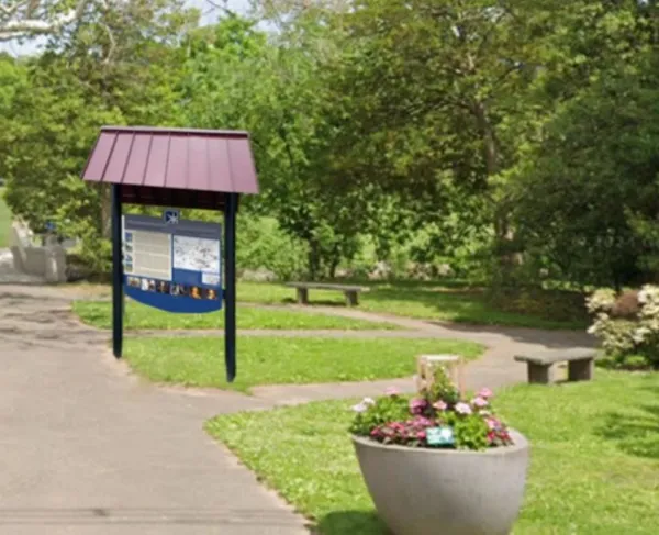 A red‑roofed informational sign stands beside a split paved walkway in a green park, with flowers, trees, and a small bridge in the background.