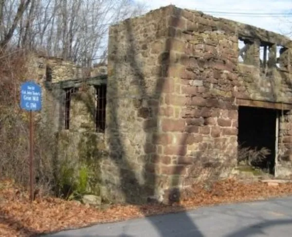 The stone ruins of an old grist mill stand among leafless trees, with a blue historical marker identifying the site beside the weathered walls.